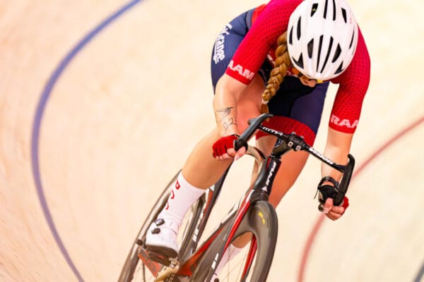 A cyclist in a red and blue uniform and white helmet leans into a turn while racing on an indoor velodrome track. The cyclist is riding a black track bike and the background shows the banked wooden surface with blue and red lines.